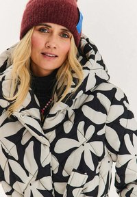 Prošívaná bunda s černobílým květinovým vzorem, vybavená vysokým límcem, předními knoflíky a měkkou texturou. Nošena s bordó pletenou čepicí.