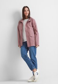 Woman leaning against wall wearing a mauve hooded jacket, light gray sweater, blue jeans, and white sneakers with dark blue accents.
