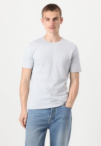 Svetlo sivo kratek rokav T-shirt iz bombaža z okroglo izrezom in majhno oznako na levem prsnem delu. V kombinaciji z modrimi džinsami.