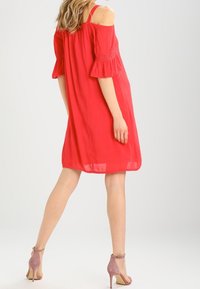 Robe rouge décolletée avec des manches courtes à volants, fabriquée en tissu léger. Complétée par des chaussures à talons roses pâles.