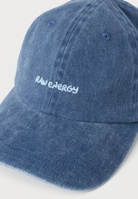 Gorra de béisbol de mezclilla en azul oscuro con visera curva y el logo bordado "RAW ENERGY" en blanco en el frente. Textura suave, diseño casual.