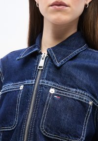 Γυναίκα που φοράει σκούρο μπλε τζιν μπουφάν με ασημένιο φερμουάρ και δύο τσέπες στο στήθος, η μία διαθέτει ένα μικρό λογότυπο με κόκκινη, λευκή και μπλε σημαία.