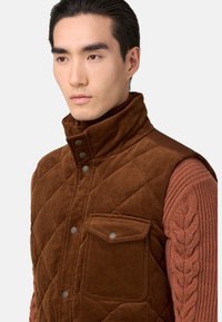 Gilet in corduroy marrone con trapuntatura a rombi, colletto alto, bottoni a pressione e una tasca frontale. Indossato sopra un maglione rosso textured.