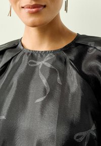 Juoda bluzonė su blizgiu paviršiumi, turinti subtilius sidabrinius taškuotus lankus ir surinkimus ant iškirptės, minkšta tekstūra.