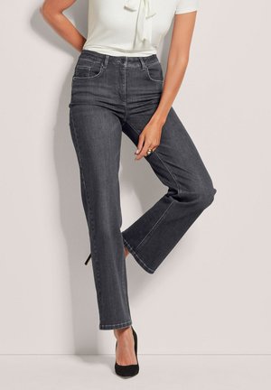Femme portant un jean gris foncé taille haute, jambes droites, un haut blanc à manches courtes avec un nœud, des talons noirs, main reposant sur la cuisse.