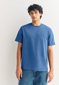 Blaues Baumwoll-T-Shirt mit kurzen Ärmeln und rundem Ausschnitt, mit glatter Textur und entspanntem Schnitt. Kombiniert mit Jeans-Shorts.