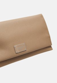 Beige kabelka na ruku vyrobená z texturovaného materiálu, s kovovou logem v stříbrné barvě a designem s přehybným uzavřením. Minimalistický a funkční vzhled.