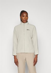 Giacca in fleece beige chiaro con colletto alto, zip frontale completa e logo sul lato sinistro del petto. Presenta maniche lunghe e una texture morbida.