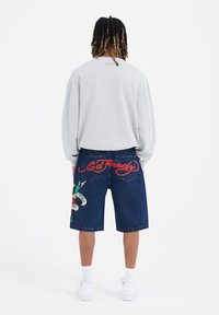 Sudadera gris claro con cuello redondo, combinada con pantalones cortos de mezclilla azul oscuro que presentan un gráfico colorido y un bordado llamativo "Ed Hardy".