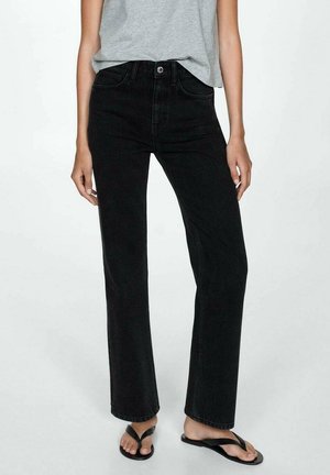 Kvinde iført sorte, højtaljede, strømpeben jeans med lige ben, grå ærmeløs top og sorte klip-klap sandaler foran en ensartet lys baggrund.