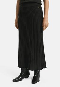 Zwarte geplooide maxi-jurk van ribstof met een aansluitende taille. Kenmerkt zich door een gouden embleemdetail aan de zijkant. Draagt met glanzende zwarte enkellaarsjes.