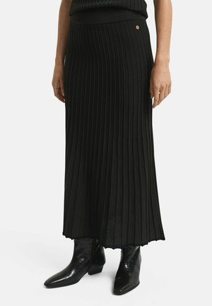 Zwarte geplooide maxi-jurk van ribstof met een aansluitende taille. Kenmerkt zich door een gouden embleemdetail aan de zijkant. Draagt met glanzende zwarte enkellaarsjes.