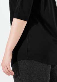 Schwarzes, kurzärmliges Oberteil mit abgerundetem Saum. Kombiniert mit schwarzen, strukturierten Hosen, die einen dezenten Glanz und ein metallisches Muster aufweisen.
