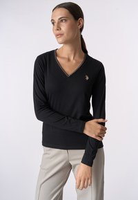 Maglietta nera a maniche lunghe con scollo a V in tessuto morbido, con un colletto a contrasto e logo ricamato sul petto. Abbinata a pantaloni chiari.
