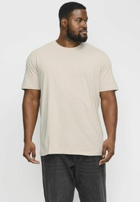 T-shirt court beige clair à manches courtes dans une coupe décontractée, fabriqué en tissu doux. Associé à un pantalon gris foncé, porté par une personne de profile.