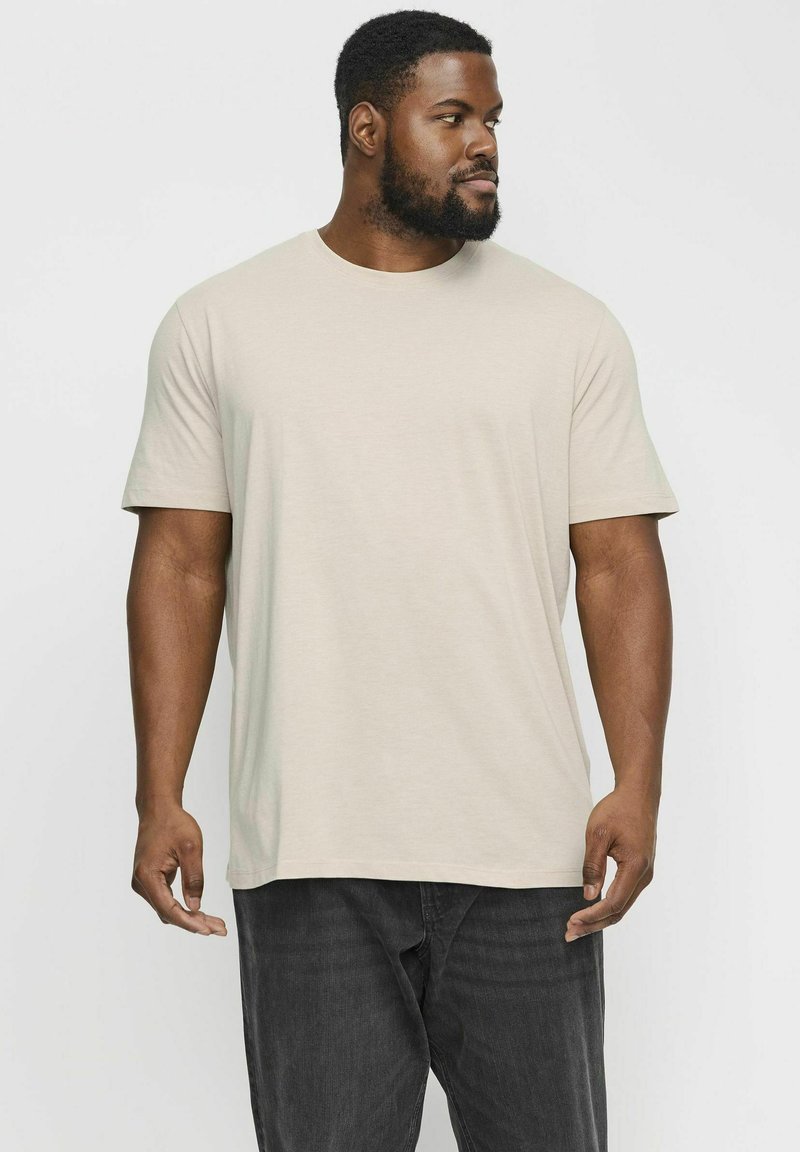 T-shirt court beige clair à manches courtes dans une coupe décontractée, fabriqué en tissu doux. Associé à un pantalon gris foncé, porté par une personne de profile.