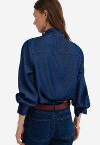 Donkerblauwe denim overhemd met een ontspannen pasvorm, kraag en knoopmanchetten. Bevat een rugpand en is gecombineerd met donkerblauwe jeans.
