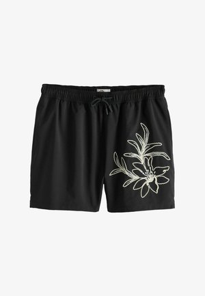 Prim-plan al taliei unei persoane care poartă pantaloni scurți negri cu talie elastică, șnur negru și design brodat cu frunze de culoare crem.