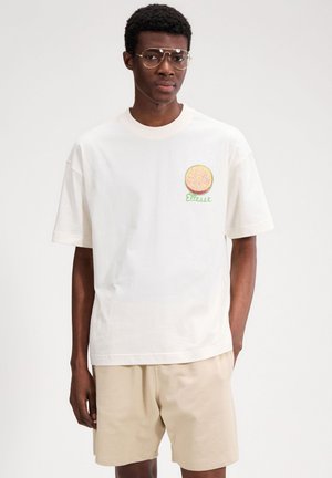 Mann mit runder Brille, weißem T-Shirt mit Zitrusfrucht-Logo und dem Schriftzug "Ellesse", beigen Shorts, steht vor einfarbigem Hintergrund.