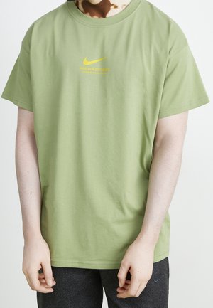 Osoba ubrana w jasnozielony t-shirt Nike Athletic Dept z żółtym logo i ciemnoszare spodnie Nike, ramiona rozluźnione wzdłuż ciała.