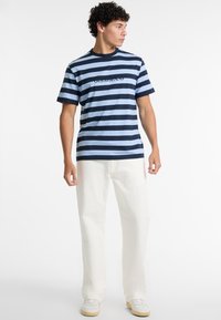T-shirt bleu et navy à rayures avec des manches courtes et un col rond, arborant un graphique "GUESS JEANS" au centre. Porté avec un pantalon blanc.