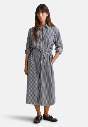 Frau trägt ein schwarz-weiß vertikal gestreiftes midi Hemdblusenkleid mit hochgekrempelten Ärmeln, gebundenem Taillengürtel und schwarzen Schnürschuhen, stehend.
