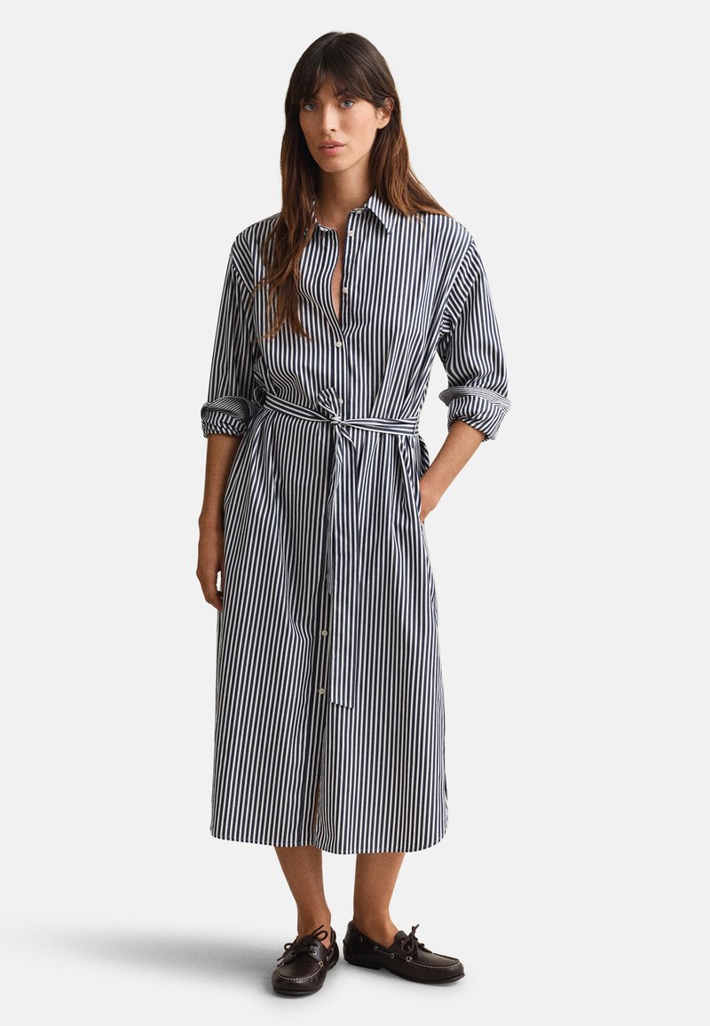 Γυναίκα που φοράει μακρυμάνικο midi πουκάμισο-φόρεμα με κάθετες μαύρο-λευκές ρίγες, με ρεβέρ στα μανίκια, ζώνη στη μέση που δένει, και μαύρα δερμάτινα παπούτσια με κορδόνια, όρθια.