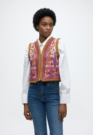 Donna che indossa una camicia bianca a maniche lunghe, jeans blu e un gilet ricamato colorato con motivi floreali e rifiniture marroni.