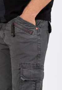 Graue Cargo-Hosen verfügen über eine strukturierte Oberfläche, seitliche Taschen mit Reißverschlüssen, rote Akzente und einen Knopfverschluss mit einem Logo.