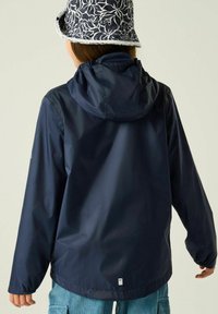 Persona que lleva una chaqueta impermeable azul marino con capucha y un sombrero tipo bucket azul con estampado, vista desde atrás sobre un fondo liso.