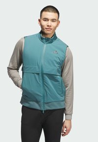 Veste sans manches teal avec fermeture éclair et poches, portée par-dessus un haut à manches longues gris clair. Présente une texture lisse.