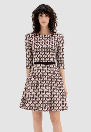 Donna con capelli ricci e scuri che indossa un vestito al ginocchio con maniche a tre quarti, un motivo geometrico e una cintura nera in vita.