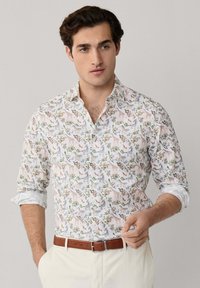 Mladý muž nosiaci bielu košeľu na gombíky s vyhrnutými rukávmi, na ktorej sú farebné potlače zvierat a rastlín, v kombinácii s béžovými nohavicami a hnedým opaskom.