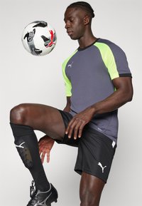 Grå og neongrønn sportskjorte med korte ermer, svarte shorts, sorte sokker og fotballsko. En svart-hvitt fotball er balansert på et kne.