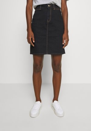 Person iført en sort denim nederdel til knæet, hvide sneakers og en stribet indstukket skjorte, stående med armene afslappet langs siden.