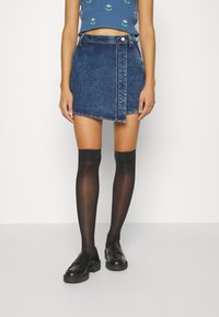 Kvinna klädd i en asymmetrisk mörk denimminikjol, svarta genomskinliga knästrumpor, svarta loafers och en blå kortärmad topp med blommönster.