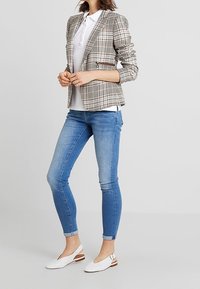 Blazer à motif prince de Galles beige et marron, associé à un polo blanc, un jean skinny bleu et des chaussures blanches ouvertes avec des accents marron.