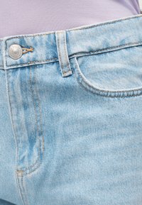 Φαρδύ γαλάζιο τζιν στη μέση με κουμπί και θηλιά για ζώνη, φορεμένο πάνω από ένα λιλά πουκάμισο, που δείχνει το εμπρόσθιο τσεπάκι και τις ραφές.