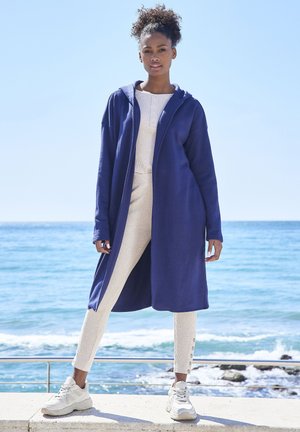 Lange marineblauwe hoodiejas van fleece, gecombineerd met lichtbeige gestructureerde broek met zijknopen en witte sneakers. Oceaan op de achtergrond.