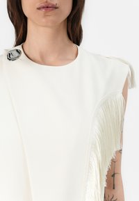 Asymmetrical white top featuring a silver accent button and long, textured fringe detail along one side. Smooth fabric and clean lines.