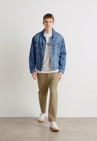Denim jaka ar divām priekšējām kabatām, valkāta virs baltas T-krekla, kombinēta ar bēšiem biksēm un baltām sporta kedām. Modelis stāv uz vienkāršas fona.