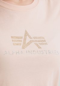 Světle růžové tričko vyrobené z měkké látky s logem z zlatých a stříbrných kamínků, které obsahuje geometrické tvary a nápis "ALPHA INDUSTRIES."