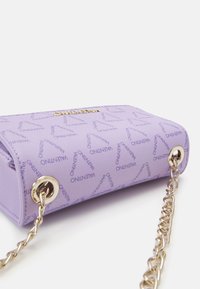 Lila handväska med texturerad tyg som har ett repetitivt logomönster, guldfärgad kedjehandtag och en rundad rektangulär form.