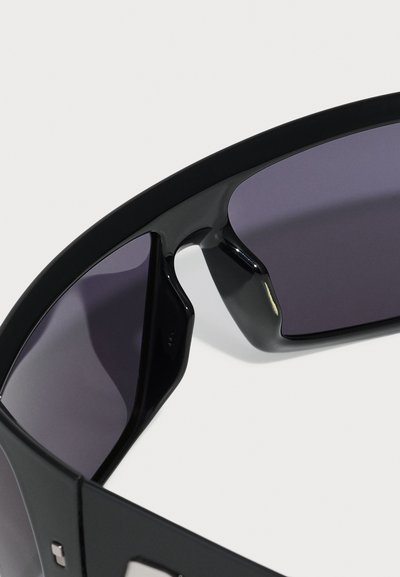 Ochelari de soare negri cu un design elegant și unghiular, având lentile întunecate și ramă din plastic netedă. Un detaliu notabil include un accent argintiu.