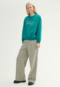 Zielona bluza z nadrukiem "COAST" w białym kolorze, połączona z beżowymi szerokimi spodniami w kratę oraz czarnymi butami na platformie. Gładka faktura.
