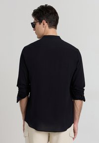 Schwarzes Shirt aus strukturiertem Stoff mit einem klassischen Kragen und hochgekrempelten Ärmeln. Die Rückseite ist glatt und weist subtile Nähte auf.