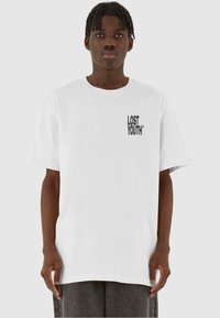 Balta medvilninė marškinėlių su juoda spauda "LOST YOUTH" logotipo kairėje krūtinės pusėje. Paprastas dizainas, laisvas kirpimas ir trumpomis rankovėmis.