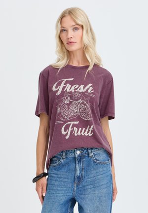 Donna bionda che indossa una t-shirt viola sbiadita con grafiche di "Fresh Fruit" e melograno, abbinata a jeans blu e un braccialetto nero.