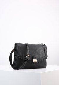 Borsa a tracolla in pelle nera con tracolla regolabile e chiusura a fibbia dorata su una superficie bianca contro uno sfondo bianco uniforme.