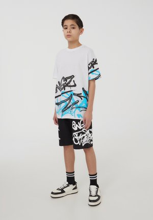 T-shirt bianca con motivi di graffiti neri e blu, abbinata a pantaloni corti neri con graffiti bianchi. Sneakers chunky in bianco e nero.